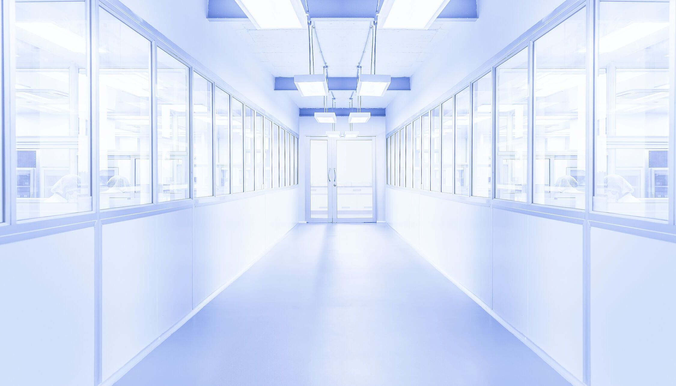 Bright white corridor with a glass door at the end. Windows on both sides reveal visible people behind them. The environment appears sterile.