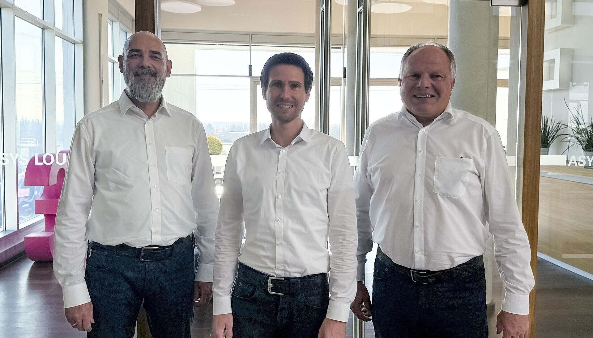 Three men wearing white shirts are standing in an office.