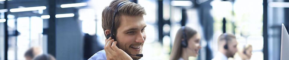 Mann mit Headset sitzt lächelnd in einem Callcenter am Schreibtisch, eine Hand am Ohr, die andere auf der Tastatur.