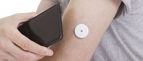 Ein schwarzes Smartphone wird an einen Arm mit Blutzuckermesser gehalten