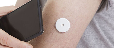 Ein schwarzes Smartphone wird an einen Arm mit Blutzuckermesser gehalten