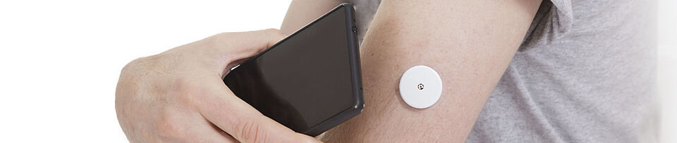 Ein schwarzes Smartphone wird an einen Arm mit Blutzuckermesser gehalten
