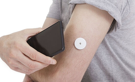 Ein schwarzes Smartphone wird an einen Arm mit Blutzuckermesser gehalten