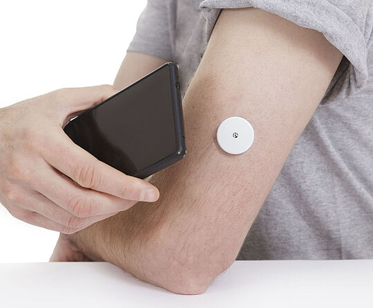 Ein schwarzes Smartphone wird an einen Arm mit Blutzuckermesser gehalten