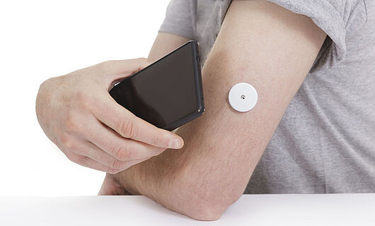 Ein schwarzes Smartphone wird an einen Arm mit Blutzuckermesser gehalten