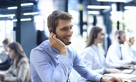 Smiling man wearing a headset sits at a desk in a call center, one hand on his ear, the other on the keyboard.