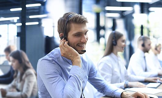 Mann mit Headset sitzt lächelnd in einem Callcenter am Schreibtisch, eine Hand am Ohr, die andere auf der Tastatur.