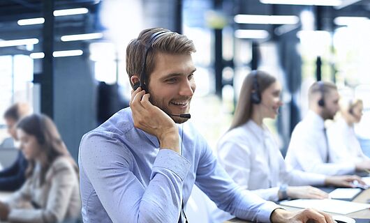 Smiling man wearing a headset sits at a desk in a call center, one hand on his ear, the other on the keyboard.