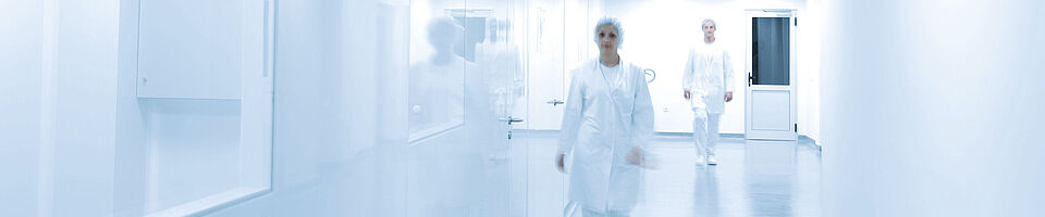 Bright white corridor with a window on the left side. A woman and a man in full laboratory clothing walk toward the camera, with the man slightly behind the woman.