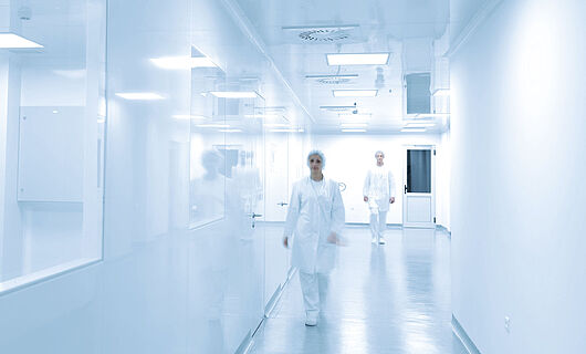 Bright white corridor with a window on the left side. A woman and a man in full laboratory clothing walk toward the camera, with the man slightly behind the woman.