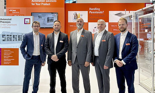 Five men in suits are standing in front of an orange exhibition stand.